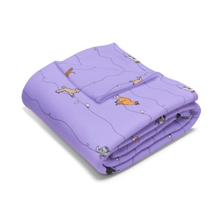 May include: Folded lavender blanket featuring a repeating pattern of cartoon dogs in various colors and poses. The dogs are connected by a wavy gray line with small white heart shapes. The blanket is folded neatly.