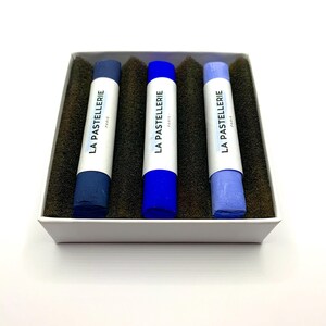 May include: A set of three blue pastel chalks in a white box with the text "LA PASTELLERIE PARIS" printed on each chalk. The chalks are shades of dark blue, blue, and light blue, resting on a dark brown foam.