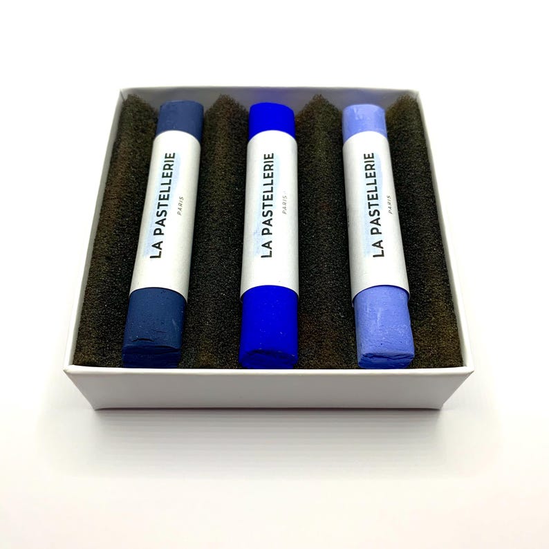 May include: A set of three blue pastel chalks in a white box with the text "LA PASTELLERIE PARIS" printed on each chalk. The chalks are shades of dark blue, blue, and light blue, resting on a dark brown foam.