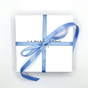 May include: A white square gift box tied with a light blue satin ribbon in a cross pattern, with a bow on top. The box has the text "LA P&Acirc;TISSERIE IS" printed on the front.