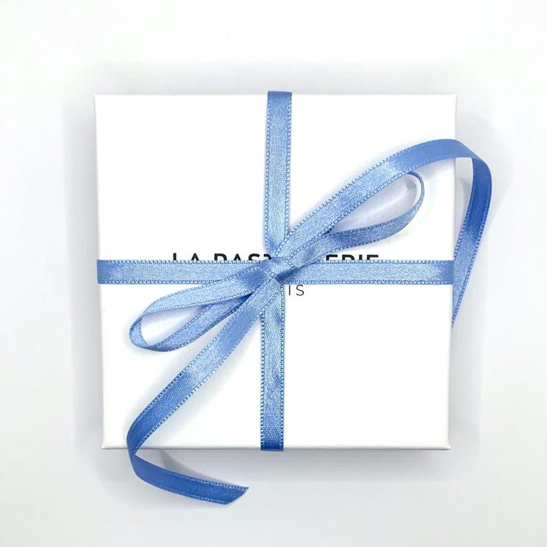 May include: A white square gift box tied with a light blue satin ribbon in a cross pattern, with a bow on top. The box has the text "LA P&Acirc;TISSERIE IS" printed on the front.