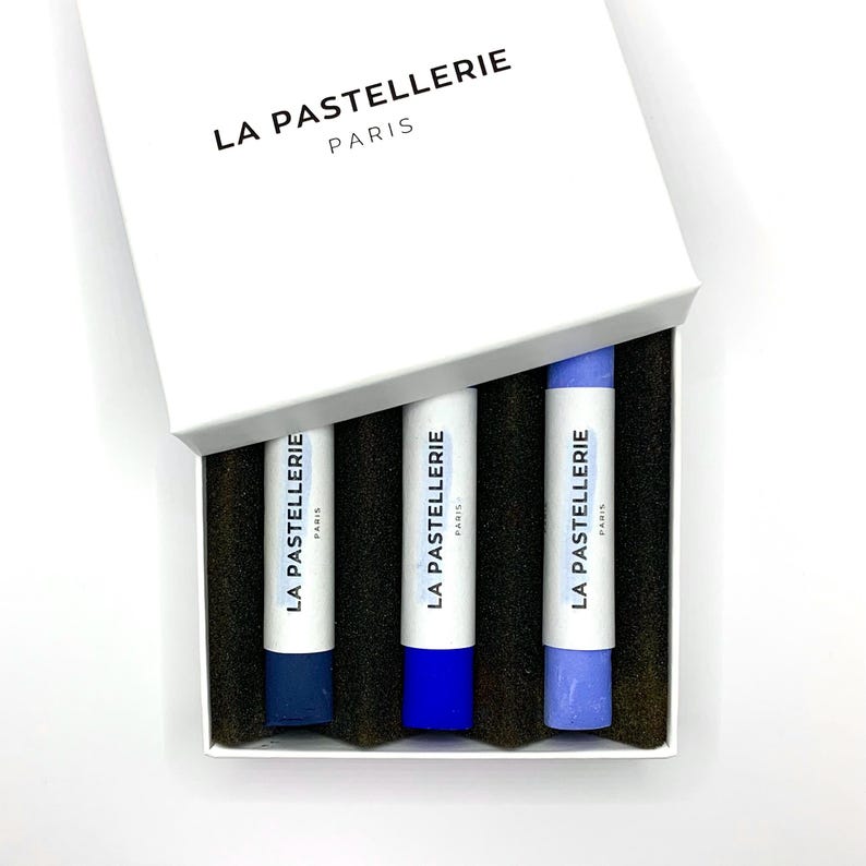 May include: A white box with the text "LA PASTELLERIE PARIS" contains three pastel sticks in shades of blue. The sticks are labeled with the same text. The box is open, revealing the contents.