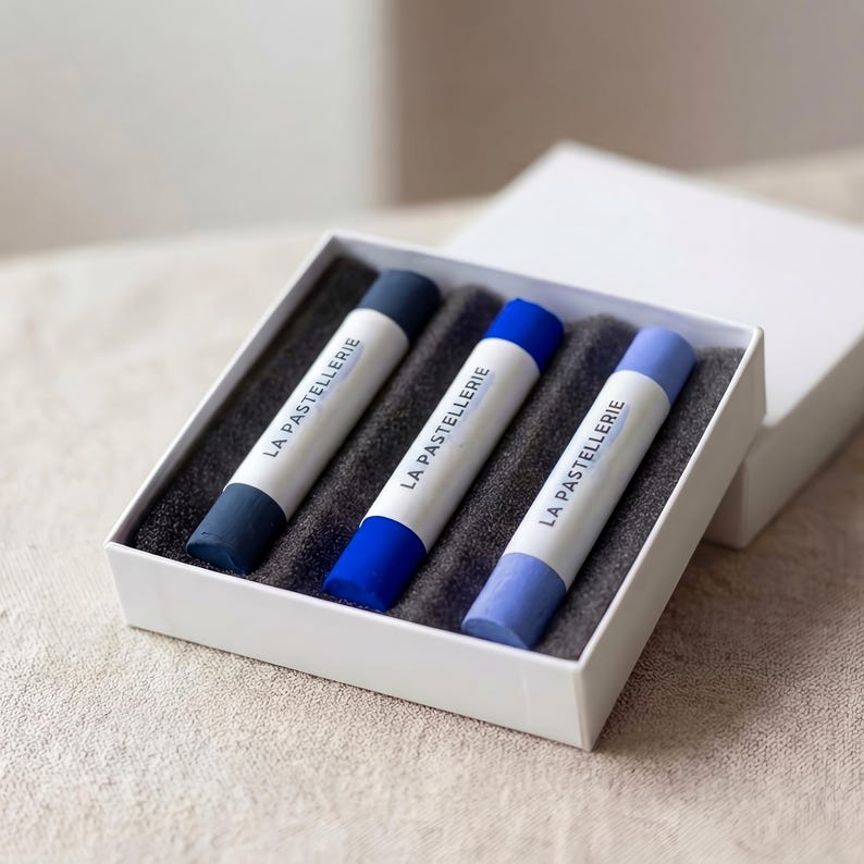 May include: A white box containing three blue pastels. Each pastel is a different shade of blue, ranging from dark to light. The pastels are labeled "LA PASTELLERIE" in a white font. The box is open, revealing the pastels nestled in a dark gray foam.