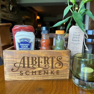 May include: A wooden condiment caddy with the words "Alberts Schenke" engraved on the front. The caddy holds four glass bottles filled with condiments, including ketchup, mayonnaise, hot sauce, and a green sauce. A green glass candle holder with a white candle is on the table next to the caddy. A sign with the text "Enjoy our fixed price lunch, Two courses £13.50" is visible in the background.