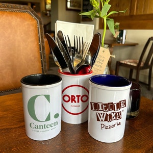 May include: Three white ceramic mugs with black, red, and blue rims. The mugs have logos for Canteen Coffee, Brunch & Bar, Orto Pizzabar, and Little Wing Pizzeria.
