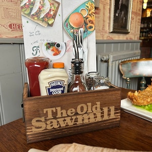 May include: A wooden condiment caddy with the text "The Old Sawmill" on the front. The caddy contains ketchup, mayonnaise, hot sauce, salt, and pepper shakers.