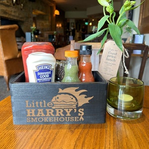 Puede incluir: Un caddy de madera para condimentos con las palabras "Little Harry's Smokehouse" en la parte delantera. El caddy contiene una botella de mayonesa Heinz, una botella de ketchup, una botella de salsa verde, una botella de salsa roja y un pequeño recipiente de granos de pimienta negra. El caddy está sentado en una mesa de madera junto a un portavelas de vidrio verde.