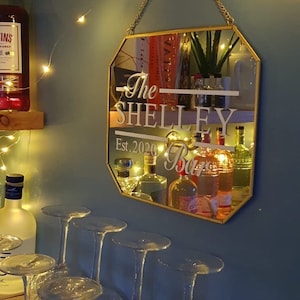 Puede incluir: Un espejo octogonal dorado con el texto "The Shelley Bar Est. 2020" colgado en una pared azul con luces de cadena. El espejo refleja una barra con botellas y vasos.