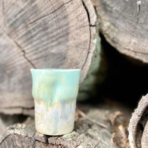 May include: A handmade ceramic tumbler with a unique, textured surface. The top half is a vibrant turquoise, transitioning to a creamy white and pale blue at the base. The cup is approximately 4 inches tall.