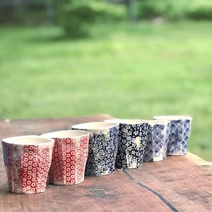 May include: A collection of six ceramic cups with unique patterns. The cups feature designs in red, white, and blue, including floral and geometric patterns. They are arranged on a wooden surface against a blurred green background.