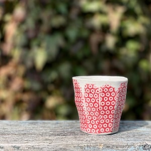 May include: A white ceramic cup with a red pattern of small circles. The cup has a slightly irregular shape and sits on a weathered wooden surface. The background is a blurred view of green foliage.
