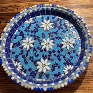 May include: A blue and white mosaic tile bird bath with a decorative design of white flowers. The bird bath is round and has a raised rim.