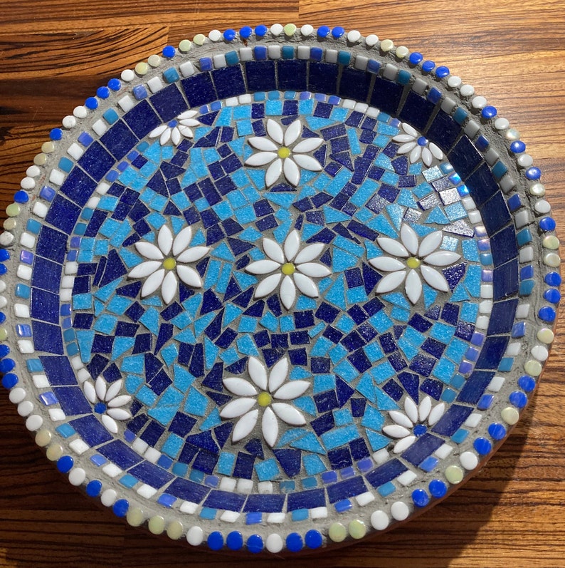 May include: A blue and white mosaic tile bird bath with a decorative design of white flowers. The bird bath is round and has a raised rim.