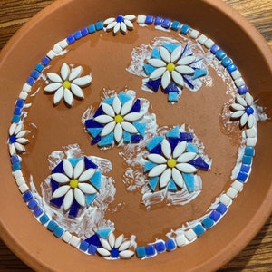 May include: A terracotta plate with a mosaic design of blue and white flowers. The flowers are made of small ceramic tiles and are arranged in a circular pattern. The plate is covered in a layer of white glue.