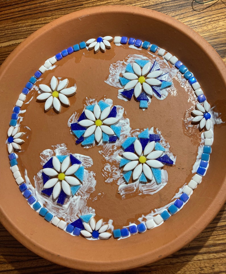 May include: A terracotta plate with a mosaic design of blue and white flowers. The flowers are made of small ceramic tiles and are arranged in a circular pattern. The plate is covered in a layer of white glue.