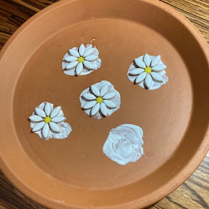 May include: A terracotta planter with four white daisy-shaped tiles with yellow centers. The tiles are arranged in a circle on the planter. There is a white blob of glue in the center of the planter.