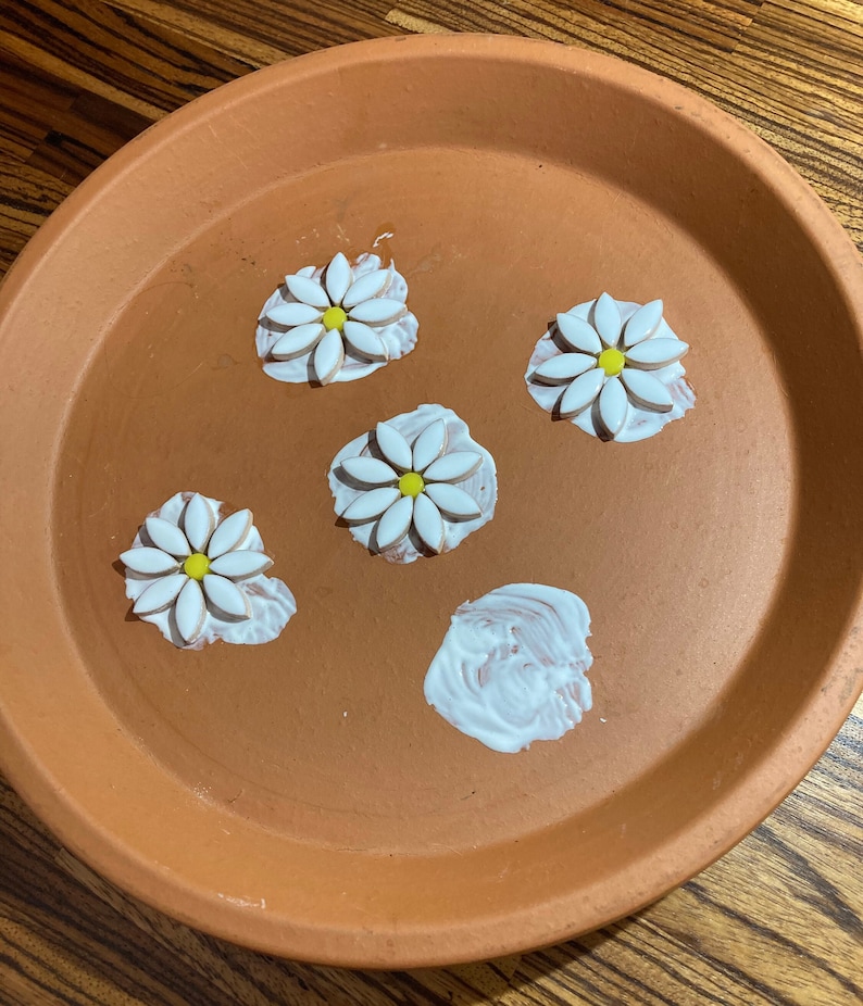 May include: A terracotta planter with four white daisy-shaped tiles with yellow centers. The tiles are arranged in a circle on the planter. There is a white blob of glue in the center of the planter.