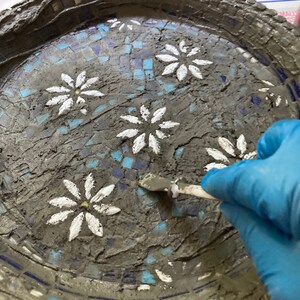 May include: A close-up of a gray concrete mosaic tray with white painted daisy flowers. A gloved hand is using a tool to smooth out the grout between the tiles.