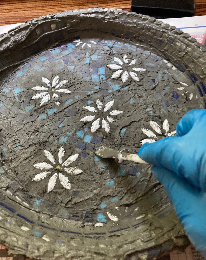 May include: A close-up of a gray concrete mosaic tray with white painted daisy flowers. A gloved hand is using a tool to smooth out the grout between the tiles.