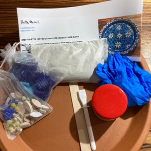 May include: A mosaic bird bath kit with a terracotta base, white grout, blue gloves, a wooden spreader, a small jar of sealant, and a bag of blue, white, and yellow mosaic tiles.