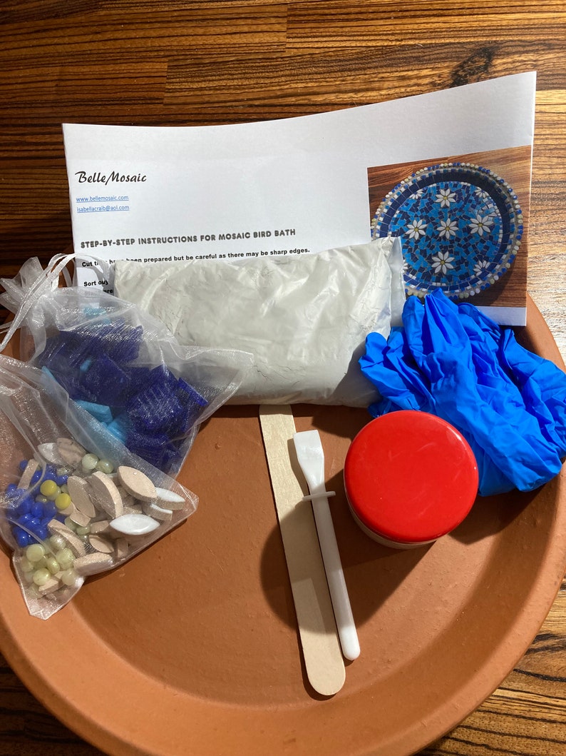 May include: A mosaic bird bath kit with a terracotta base, white grout, blue gloves, a wooden spreader, a small jar of sealant, and a bag of blue, white, and yellow mosaic tiles.