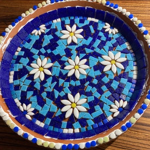 May include: A terracotta plate with a blue and turquoise mosaic design featuring white daisy flowers. The plate has a white rim with blue and yellow dots.