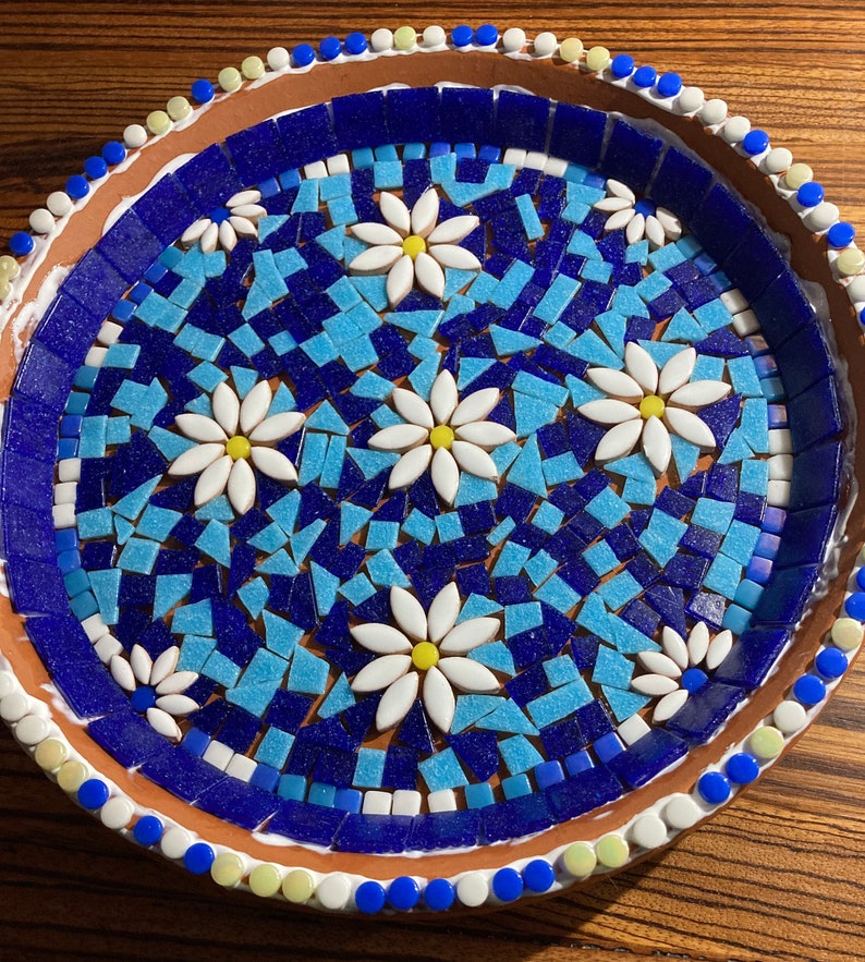 May include: A terracotta plate with a blue and turquoise mosaic design featuring white daisy flowers. The plate has a white rim with blue and yellow dots.