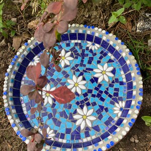 May include: A blue and white mosaic bird bath with a white daisy design. The bird bath is made of concrete and has a round shape.