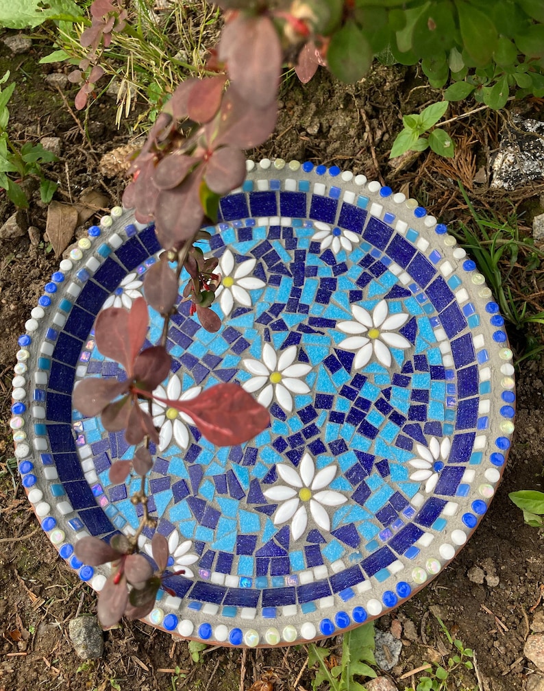 May include: A blue and white mosaic bird bath with a white daisy design. The bird bath is made of concrete and has a round shape.