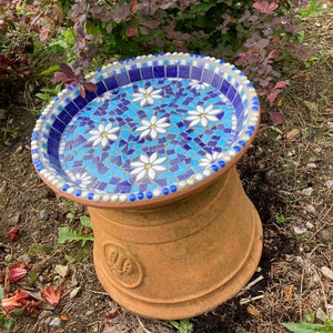 May include: A blue and white mosaic bird bath with a terracotta base. The bird bath is decorated with white flowers.
