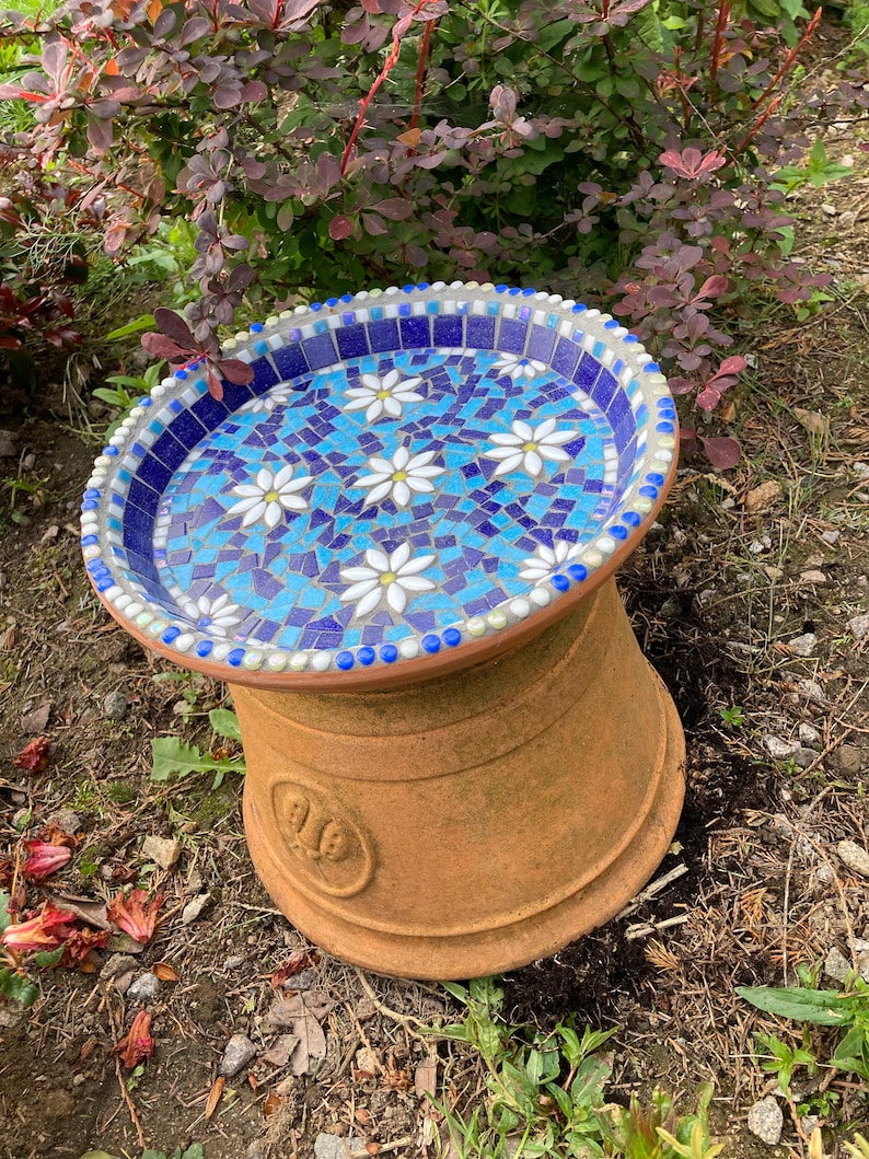 May include: A blue and white mosaic bird bath with a terracotta base. The bird bath is decorated with white flowers.