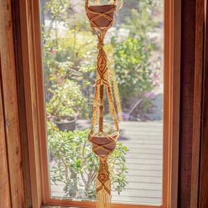 May include: A macrame plant hanger with two terracotta pots, hanging in front of a window with a view of green plants and a wooden deck.