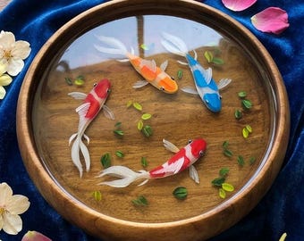 May include: A round wooden bowl filled with clear resin containing three painted koi fish in orange, blue, and red with white accents. Small green leaf-like shapes are embedded in the resin, creating a pond effect within the circular wooden frame.