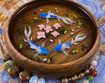 May include: A round wooden bowl containing a clear resin layer with three blue and white fish figures, several small pink flower blossoms, and scattered green leaf accents, creating a pond-like appearance within the dark wood vessel.