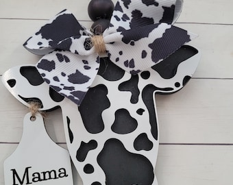 Puede incluir: Un recorte de vaca de madera con un diseño de estampado de vaca en blanco y negro. La vaca tiene un lazo blanco y negro atado alrededor del cuello y una etiqueta blanca que dice "Mama".