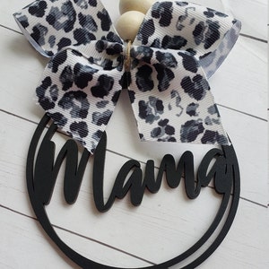May include: Black wooden circle with the word "Mama" written in a script font. A white and black leopard print bow is attached to the top of the circle with wooden beads.