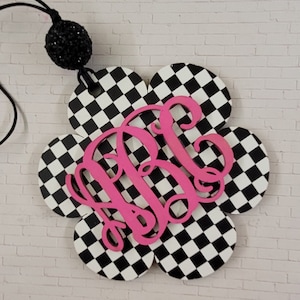 May include: A black and white checkered flower-shaped car air freshener with a pink monogram "KBC" in the center. The air freshener has a black string for hanging.