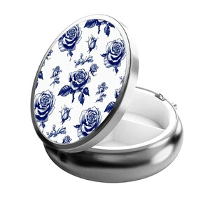 May include: A silver pill box with a white lid featuring a blue floral pattern of roses. The pill box has three compartments.