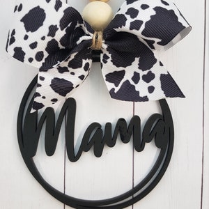 Puede incluir: Lazo de cinta de estampado de vaca blanco y negro con cuentas de madera y un círculo de madera negro con la palabra "Mama" en letra cursiva.