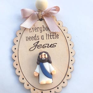 May include: Wooden oval ornament with scalloped edges, featuring the text "Everybody needs a little Jesus" and a small Jesus figurine. A pink ribbon bow and wooden bead are attached to the top with a jute string.