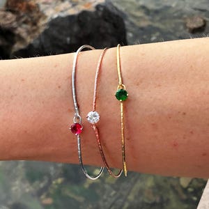 Może przedstawiać: Trzy delikatne bransoletki w kolorze srebrnym, różowego złota i złota. Każda ma pojedynczy kamień szlachetny: czerwony, przezroczysty i zielony. Bransoletki są cienkie i zakrzywione, z teksturowanym wzorem na metalu.