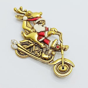 Könnte beinhalten: Eine Brosche aus goldfarbenem Metall, die den Weihnachtsmann auf einem Motorrad mit einem Rentier auf dem Rücksitz zeigt. Das Design besticht durch rote und weiße Emaille-Akzente am Anzug des Weihnachtsmanns und zwei klare, marquise-geschliffene Kristalle an den Auspuffrohren.