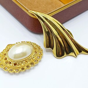 Könnte beinhalten: Eine Brosche aus goldfarbenem Metall mit einer glatten, geschwungenen, blattartigen Form neben einer zweiten Brosche, die aus einer ovalen Kunstperle in einem strukturierten, durchbrochenen goldfarbenen Metallrahmen besteht.