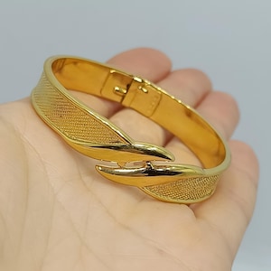 Könnte beinhalten: Ein goldfarbener Scharnierarmreif mit polierter Oberfläche und texturierten, kreuzschraffierten Details auf der Vorderseite. Das Armband hat ein geschwungenes, überlappendes Design in Blattform und wird mit einem Metallverschluss gesichert.