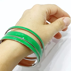 Puede incluir: Tres pulseras de esmalte verde con ribete plateado apiladas en una muñeca. El color verde brillante de las pulseras contrasta con el ribete plateado, creando un efecto visual llamativo. Las pulseras son un accesorio sencillo pero elegante.