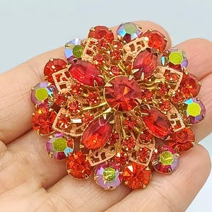 Puede incluir: Broche de estilo vintage con diseño floral. La base es de metal dorado y está adornada con pedrería roja e iridiscente de varios tamaños. La piedra central es una gema roja grande y facetada.