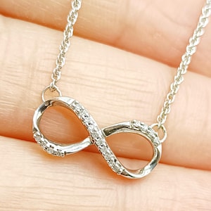 Puede incluir: Un collar con colgante de símbolo de infinito en tono plateado que presenta una cadena delicada y un dije de bucle de infinito metálico. La superficie del dije está adornada con pequeñas piedras claras y brillantes a lo largo del marco.
