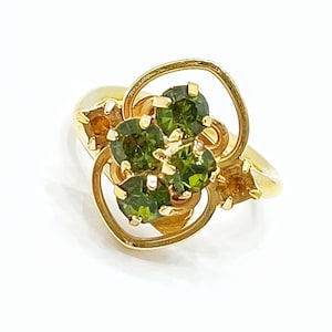 May include: A gold-toned ring featuring a central cluster of four round, deep green faceted stones held in prong settings. The ring is accented by two smaller, amber-coloured stones on the sides, all framed within a swirling, open-work gold metal design.