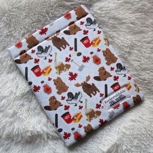 Puede incluir: Una bolsa de tela blanca con un estampado de temática canadiense que presenta castores, alces, hojas de arce y tazas de café Tim Hortons. La bolsa tiene una etiqueta que dice "Booknerdchicks".