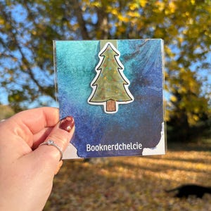 Könnte beinhalten: Ein Lesezeichen mit einem Weihnachtsbaum mit grünem Baumdesign und bunten Lichtern. Das Lesezeichen befindet sich auf einem blau-türkisfarbenen Aquarellhintergrund. Der Text "Booknerdchelcie" ist sichtbar.